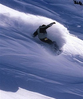 A snowboarder rides down a steep slope, lifting a cloud of white powder