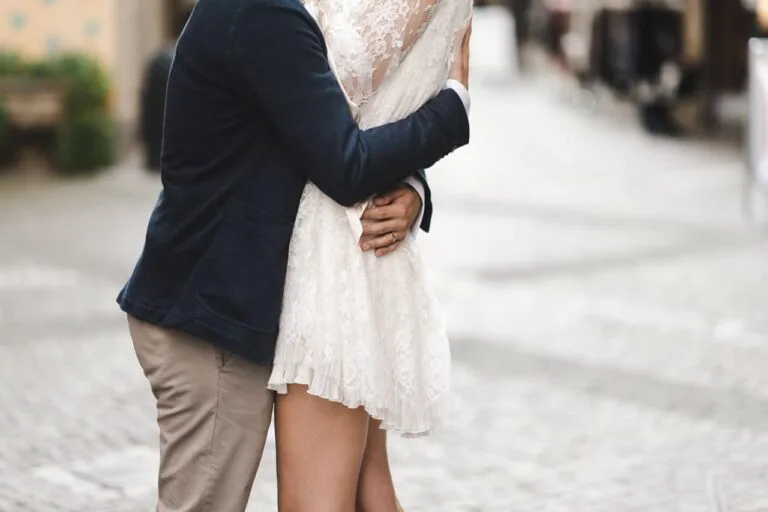 Un homme en veste bleue et pantalon beige enlace une femme en robe blanche